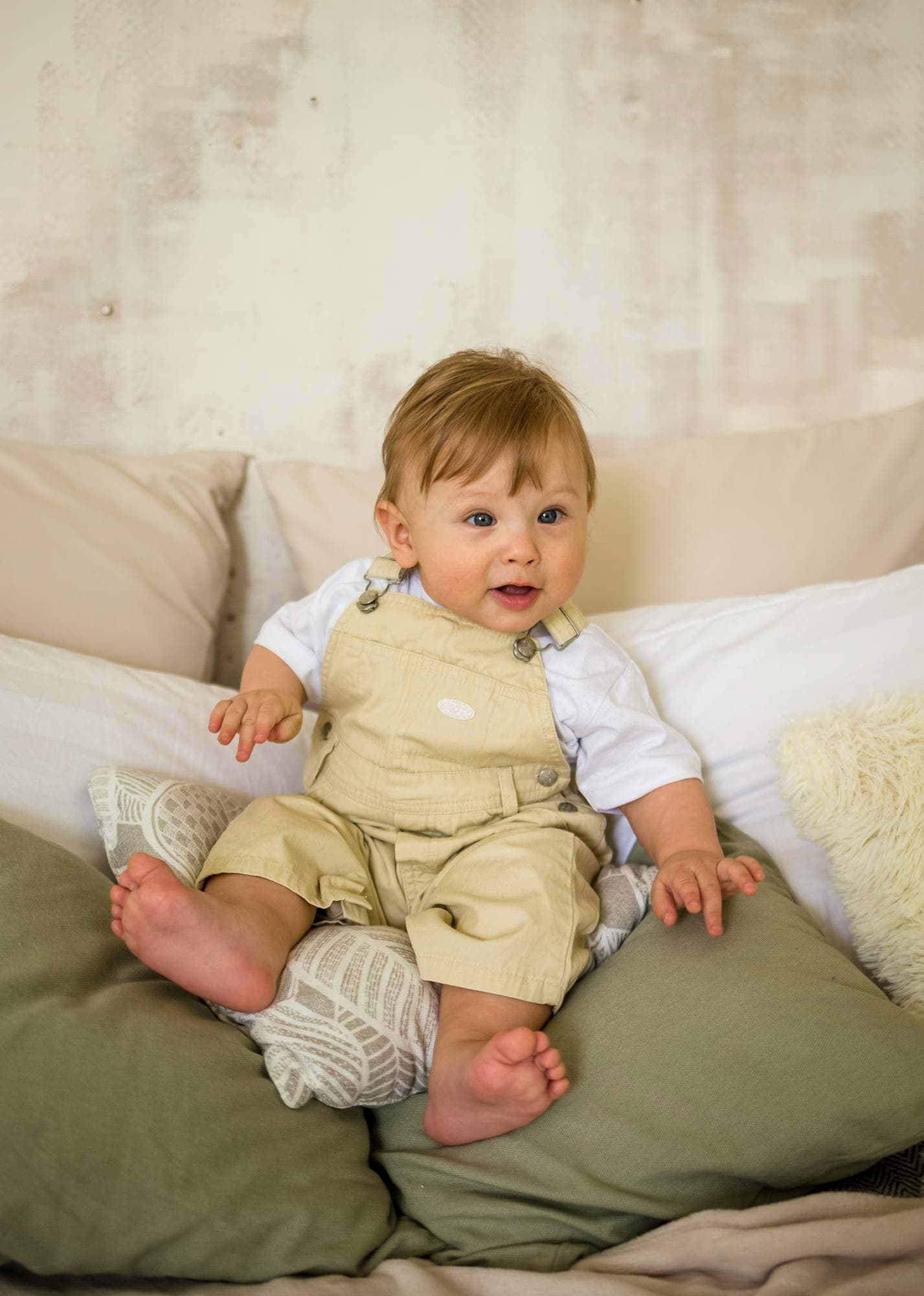 Smiling baby boy in olive Meadow dungarees on a sofa